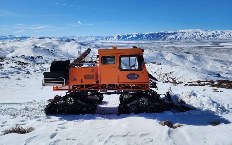 High Desert Internet's orange snowcat on a snowy summit in the Singatse Range, with the valley and snow-covered mountains behind